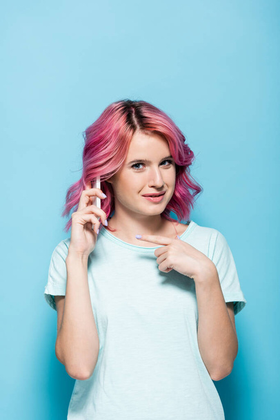 mujer joven con el pelo rosa apuntando al teléfono inteligente sobre fondo azul - Foto, Imagen