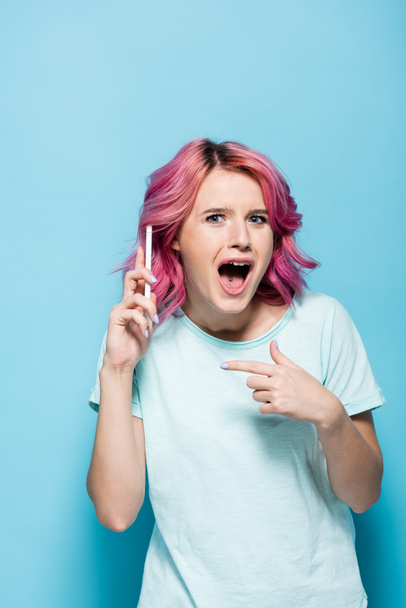 überraschte junge Frau mit pinkfarbenen Haaren zeigt auf Smartphone auf blauem Hintergrund - Foto, Bild