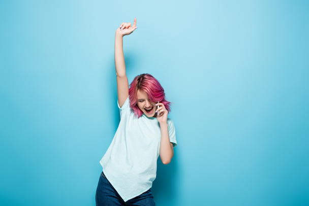 excited young woman with pink hair talking on smartphone with raised hand on blue background - Photo, Image