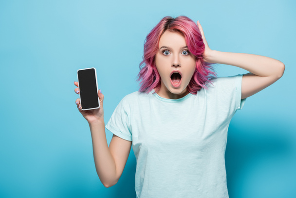 sorprendió a mujer joven con el pelo rosa sosteniendo teléfono inteligente con pantalla en blanco sobre fondo azul - Foto, imagen