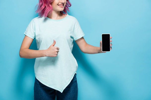 vista ritagliata di giovane donna con capelli rosa smartphone con schermo bianco e mostrando pollice su sfondo blu - Foto, immagini