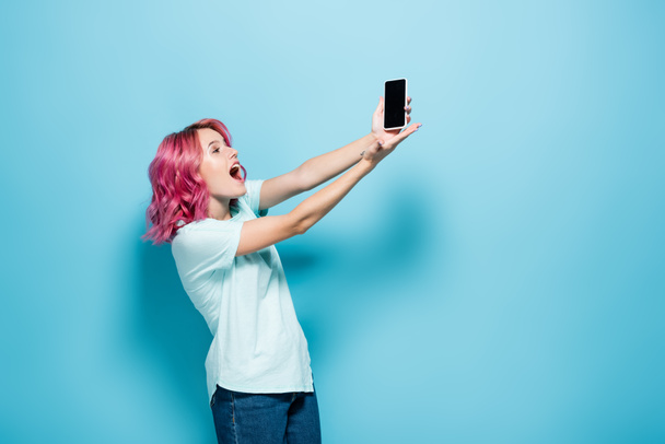 surprised young woman with pink hair holding smartphone with blank screen on blue background - Фото, изображение