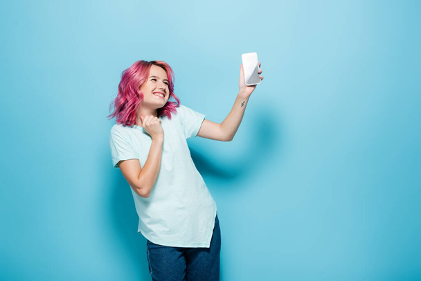 mujer joven con pelo rosa sosteniendo teléfono inteligente sobre fondo azul - Foto, imagen
