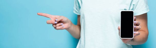 vista recortada de la mujer joven sosteniendo teléfono inteligente con pantalla en blanco y apuntando a un lado sobre fondo azul, plano panorámico - Foto, Imagen