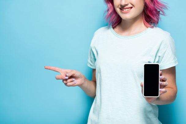 cropped view of young woman with pink hair holding smartphone with blank screen and pointing aside on blue background - Photo, image