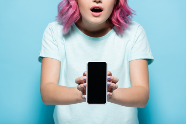 cropped view of shocked young woman with pink hair holding smartphone with blank screen on blue background - Fotó, kép