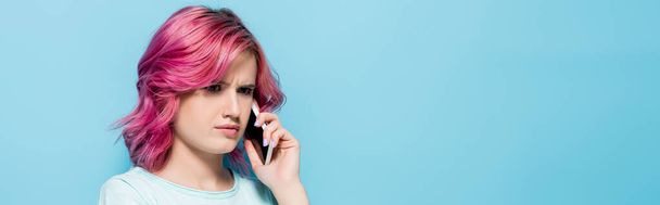 mujer joven confundida con el pelo rosa hablando en el teléfono inteligente sobre fondo azul, plano panorámico - Foto, imagen