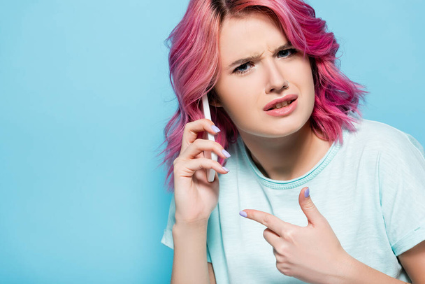 confused young woman with pink hair pointing at smartphone isolated on blue - Fotoğraf, Görsel