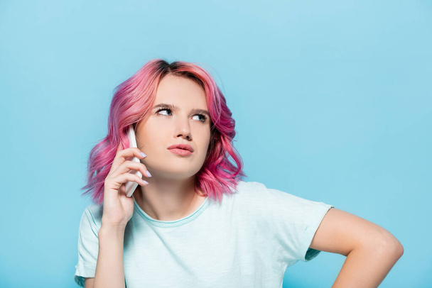 Nachdenkliche junge Frau mit pinkfarbenen Haaren spricht auf Smartphone auf blauem Hintergrund - Foto, Bild