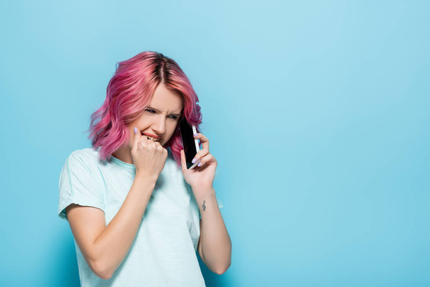 scared young woman with pink hair talking on smartphone on blue background - Fotó, kép