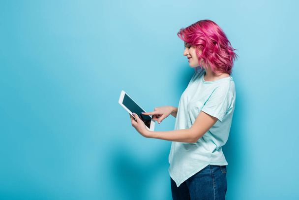 vue latérale de la jeune femme aux cheveux roses à l'aide d'une tablette numérique sur fond bleu - Photo, image