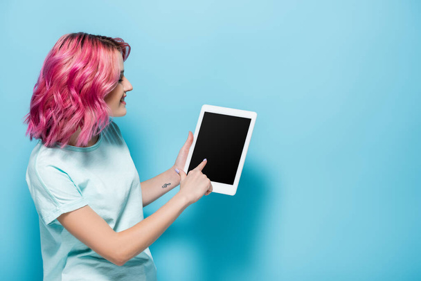 vista laterale della giovane donna con i capelli rosa utilizzando tablet digitale su sfondo blu - Foto, immagini