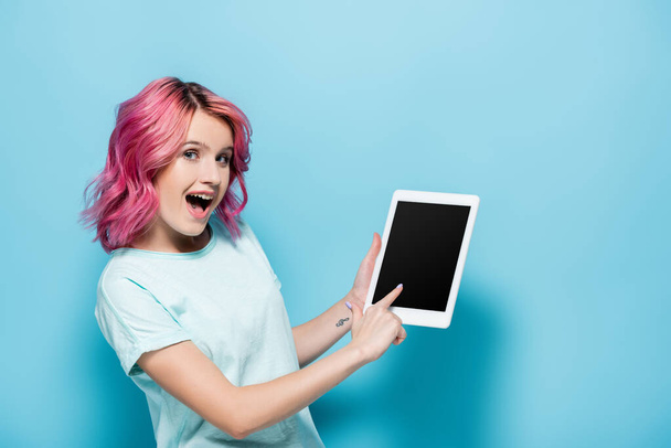 mujer joven excitada con el pelo rosa usando tableta digital sobre fondo azul - Foto, Imagen