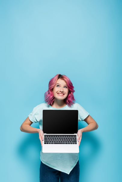 молодая женщина с розовыми волосами показывает ноутбук с чистым экраном и глядя на синий фон - Фото, изображение