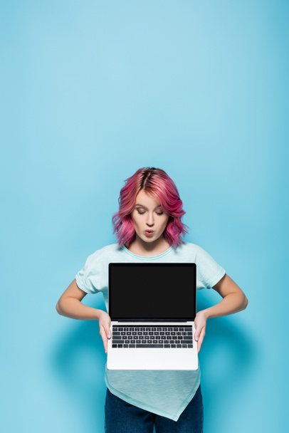 choqué jeune femme avec les cheveux roses montrant ordinateur portable avec écran blanc sur fond bleu - Photo, image