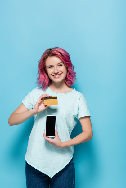 青い背景にピンクの髪をした若い女性がクレジットカードとスマートフォンを持っていて - 写真・画像