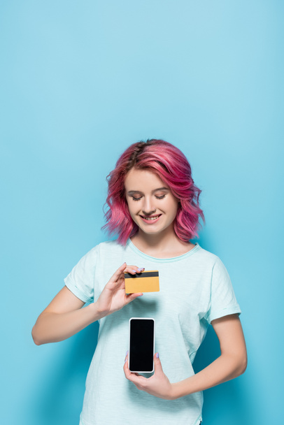 young woman with pink hair holding credit card and smartphone on blue background - Foto, afbeelding
