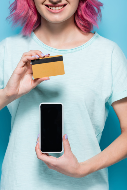 vista parcial de mujer joven con pelo rosa con tarjeta de crédito y teléfono inteligente sobre fondo azul - Foto, Imagen