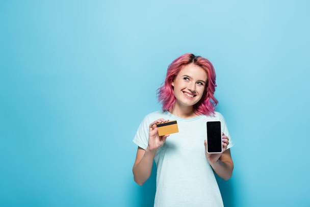 young woman with pink hair holding credit card and smartphone on blue background - Foto, Imagen