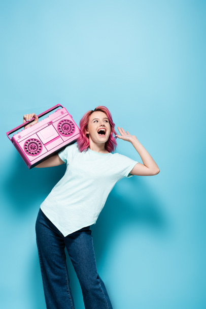 excité jeune femme avec les cheveux roses tenant magnétophone vintage sur fond bleu - Photo, image