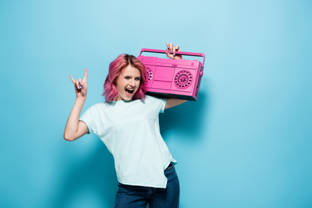 excited young woman with pink hair holding vintage tape recorder and showing rock sign on blue background - Photo, Image