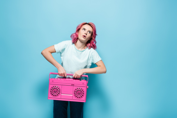 young woman with pink hair holding heavy vintage tape recorder on blue background - Photo, Image