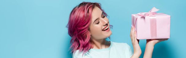 young woman with pink hair holding gift box with bow and smiling on blue background, panoramic shot - Fotografie, Obrázek