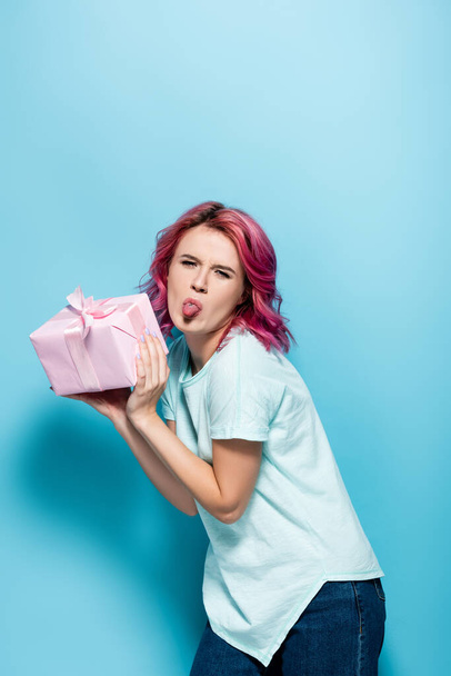 junge Frau mit rosa Haaren hält Geschenkbox mit Schleife und zeigt Zunge auf blauem Hintergrund - Foto, Bild