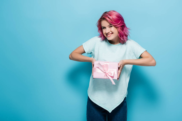 young woman with pink hair holding gift box with bow and smiling on blue background - Foto, Imagen