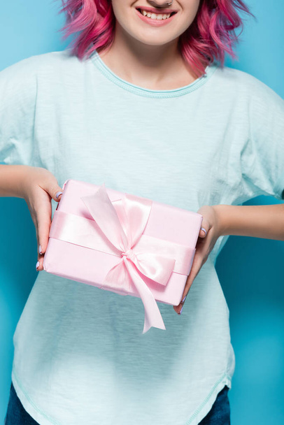 обрезанный вид молодой женщины с розовыми волосами проведение подарочную коробку с луком и улыбаясь на синем фоне - Фото, изображение