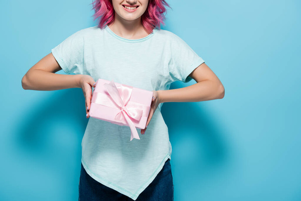 обрізаний вид молодої жінки з рожевим волоссям, що тримає подарункову коробку з бантом і посміхається на синьому фоні
 - Фото, зображення