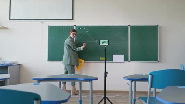 Online eğitim, karatahtanın yanında cetvelli gözlüklü pedagog okulda video dersinde cep telefonu kamerası kullanarak kendini kaydediyor. - Video, Çekim