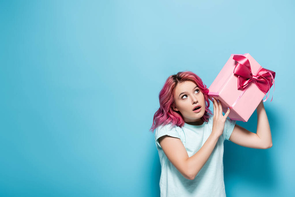 Pembe saçlı ve ağzı açık genç bir kadın mavi arka planda fiyonklu hediye kutusu tutuyor. - Fotoğraf, Görsel