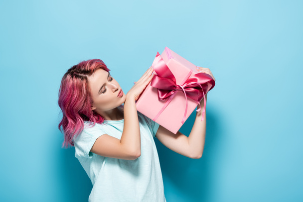 young woman with pink hair kissing gift box with bow on blue background - Photo, image