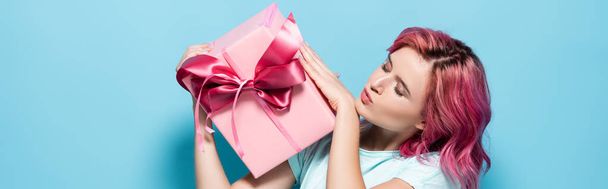 young woman with pink hair kissing gift box with bow on blue background, panoramic shot - Photo, Image