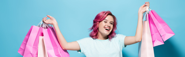young woman with pink hair holding shopping bags and smiling on blue background, panoramic shot - Photo, Image