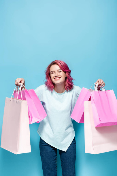 młoda kobieta z różowymi włosami trzyma torby na zakupy i uśmiecha się na niebieskim tle - Zdjęcie, obraz