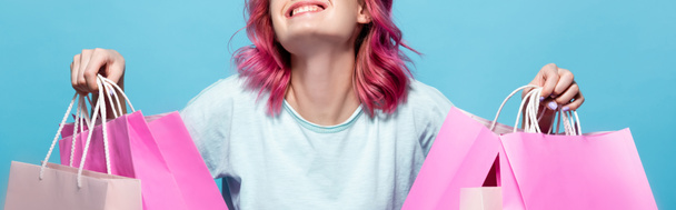 abgeschnittene Ansicht einer jungen Frau mit rosa Haaren, die Einkaufstüten in der Hand hält und isoliert auf blau lächelt - Foto, Bild