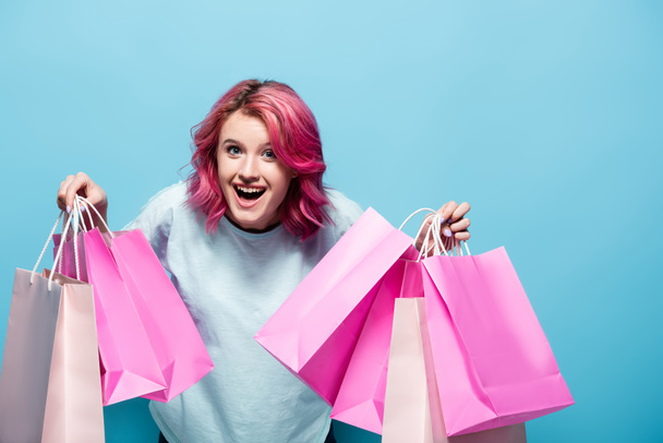excited young woman with pink hair holding shopping bags and smiling on blue background - Photo, Image