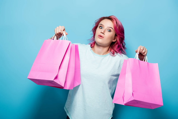 scioccato giovane donna con i capelli rosa che tiene borse della spesa su sfondo blu - Foto, immagini