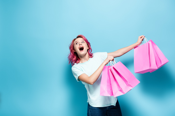 shocked young woman with pink hair holding shopping bags on blue background - Foto, immagini