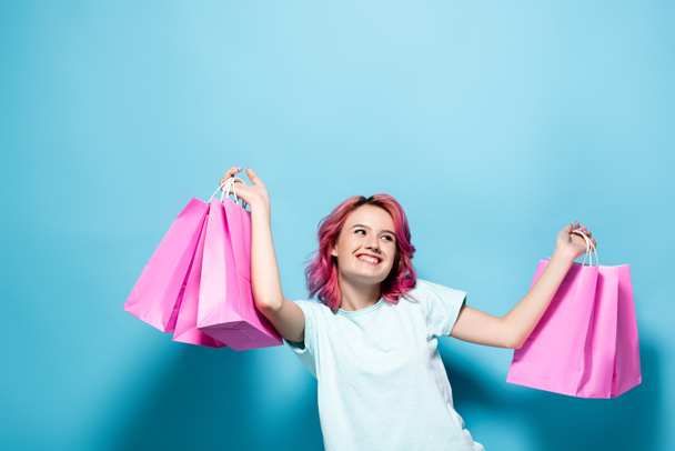 young woman with pink hair holding shopping bags and smiling on blue background - Fotoğraf, Görsel