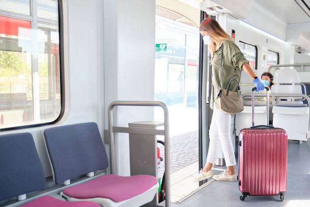 Egy fiatal nő arcmaszkban és kesztyűben, egy bőrönddel a kezében, amint leszáll a vonatról. Új normál ingázási koncepció. - Fotó, kép