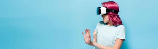 scared young woman with pink hair in vr headset gesturing on blue background, panoramic shot - Foto, imagen