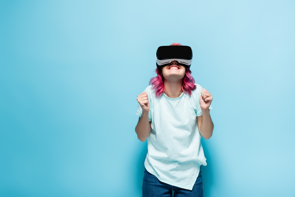 excited young woman with pink hair in vr headset showing yeah gesture on blue background - 写真・画像