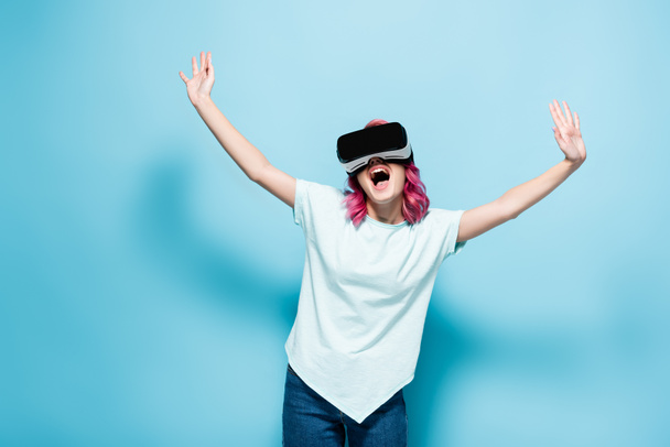 excited young woman with pink hair in vr headset with hands in air on blue background - Photo, Image