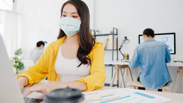 Przedsiębiorczyni z Azji, nosząca medyczną maskę na twarz, aby zachować dystans społeczny w nowej normalnej sytuacji zapobiegania wirusom podczas korzystania z laptopa w biurze. Styl życia po wirusie koronowym. - Zdjęcie, obraz