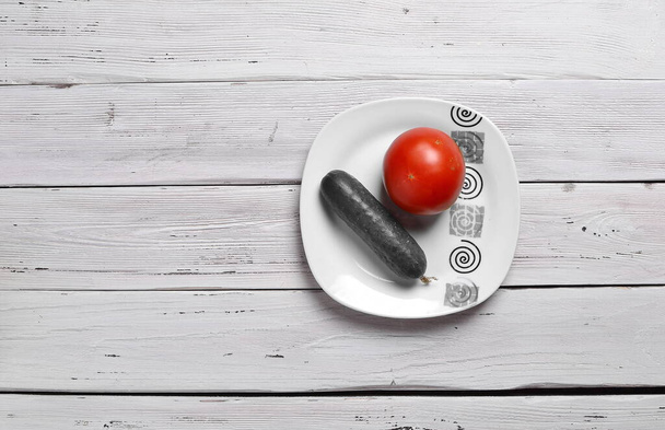 nature morte unie avec une tomate rouge et un concombre gris sur une assiette avec ornement isolé sur une vieille table rustique légère en bois - Photo, image