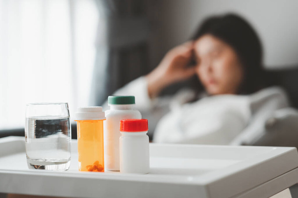 Médicaments et verre d'eau avec femme asiatique se sentant malade et maux de tête en arrière-plan dans le salon - Photo, image