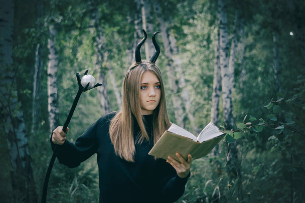 Beautiful young girl with brown hair in a witch costume, with a staff, horns, a book, against the background of a dark forest. Halloween, selective focus.Tinting for an old photo with a grainy overlay - Zdjęcie, obraz